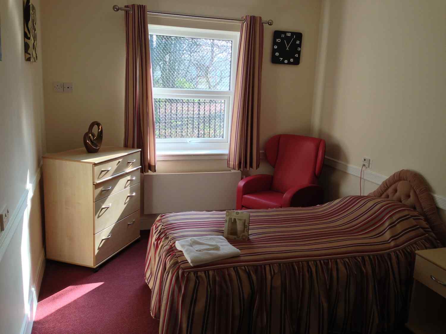 bedroom made up with red and gold bedding, a red armchair and drawers in the corner with flower wallpaper